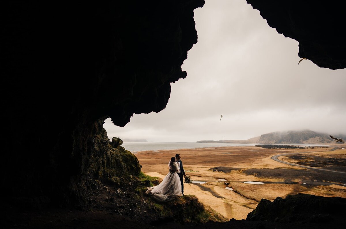 An adventurous eloping couple in Iceland, exploring caves with a breathtaking view of the ocean and mountains inside, making their journey one-of-a-kind.