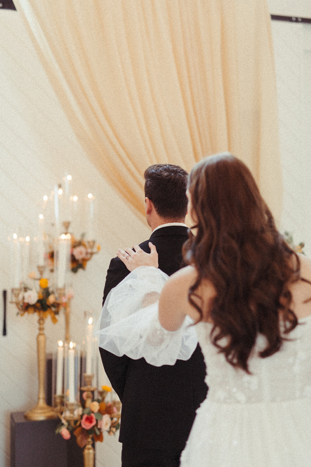 A bride wearing a white wedding gown touching the backside of the groom wearing a black tuxedo while standing at the altar.