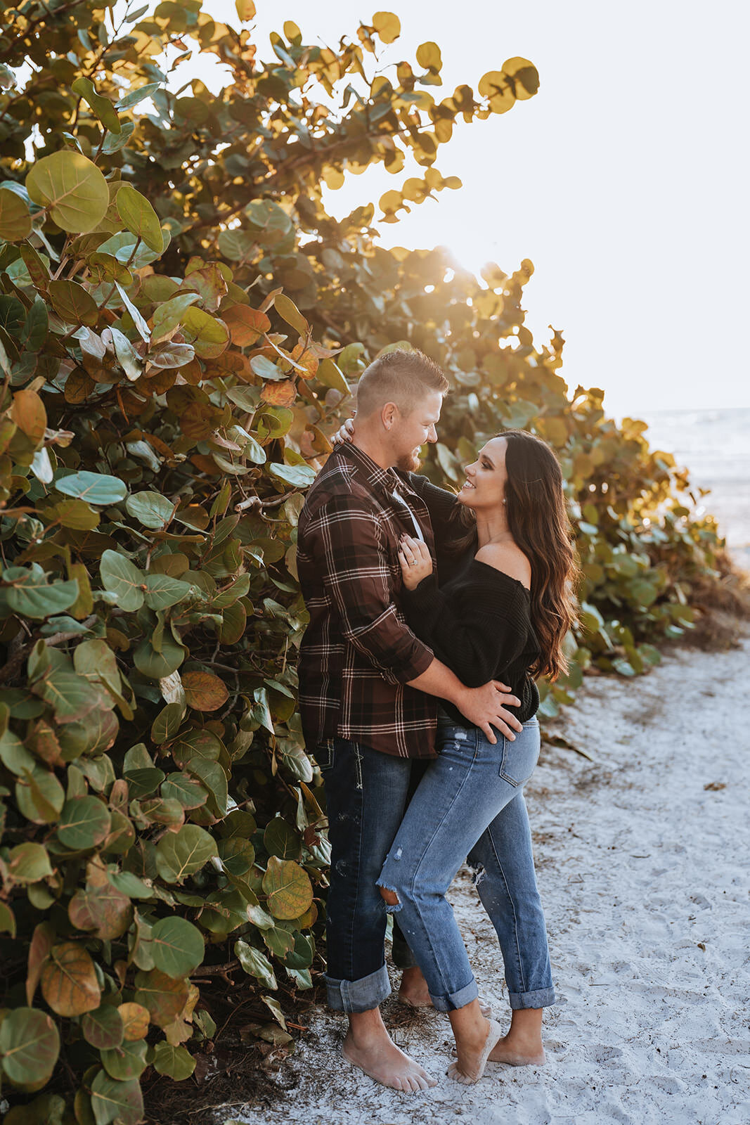couple smiling at eachother in the mangroves on the beach during engagement session wearing casual clothes including jeans