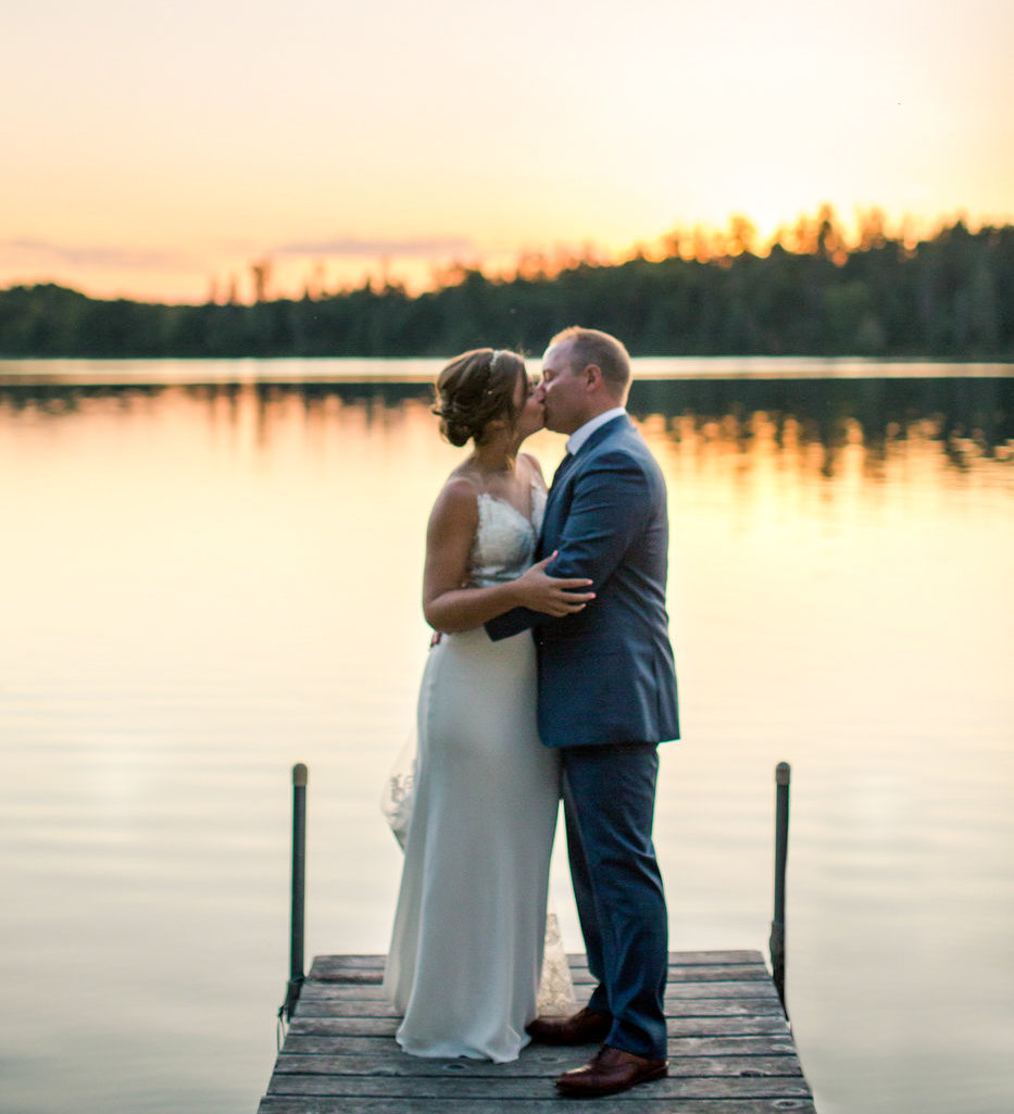 bride and groom kiss at the end of a wooden dock on lake margaret minensota at sunset