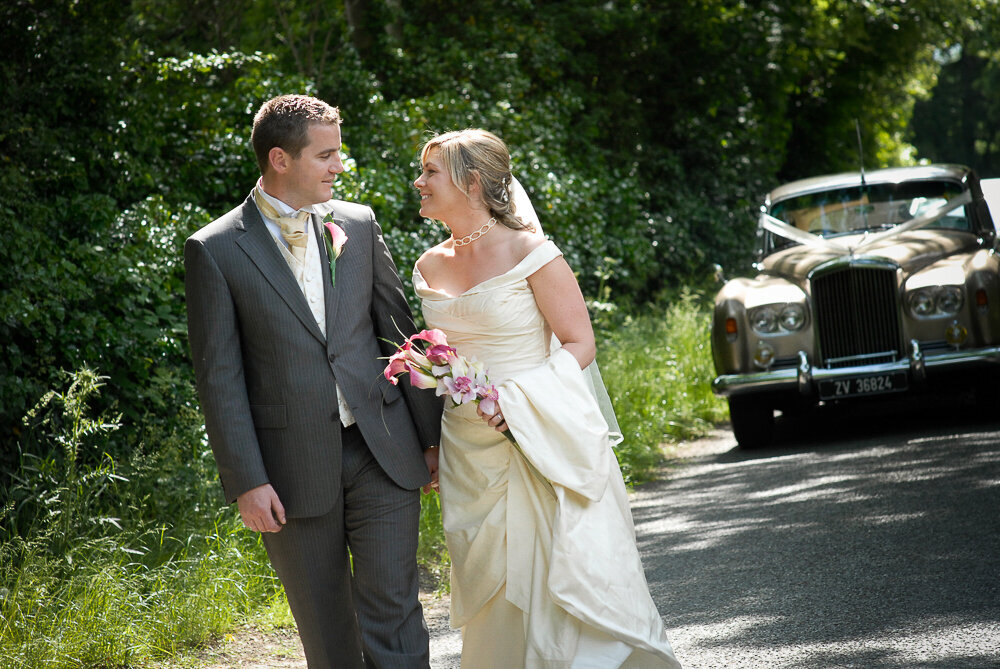 blonde bride in ivory silk walking with groom wearing a grey suit on a tree-lined country road with a vintage Rolls Royce car in the background
