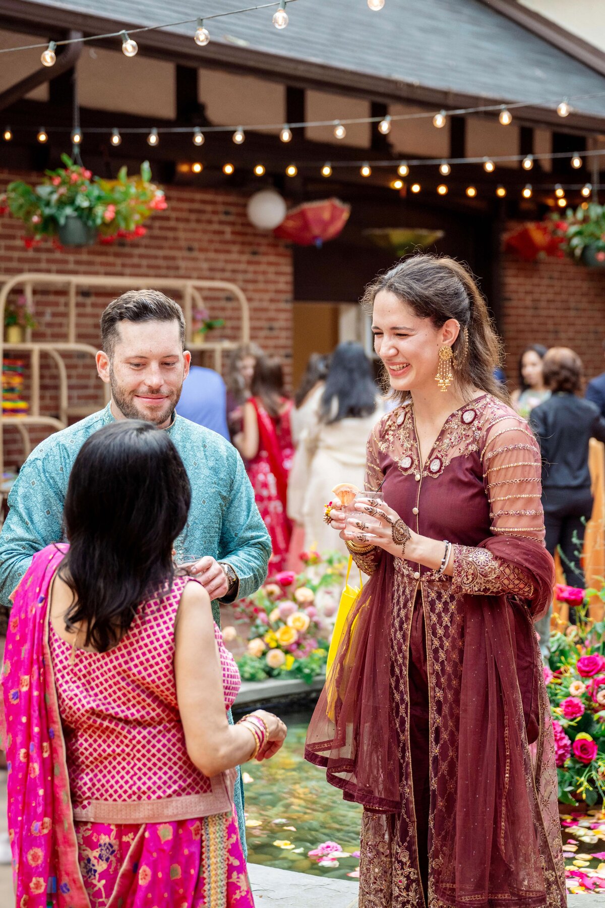 A group of people dressed in colorful traditional attire are gathered outdoors at a celebratory event. Festive decorations and string lights can be seen in the background. Everyone appears to be engaged in conversation and enjoying the occasion.