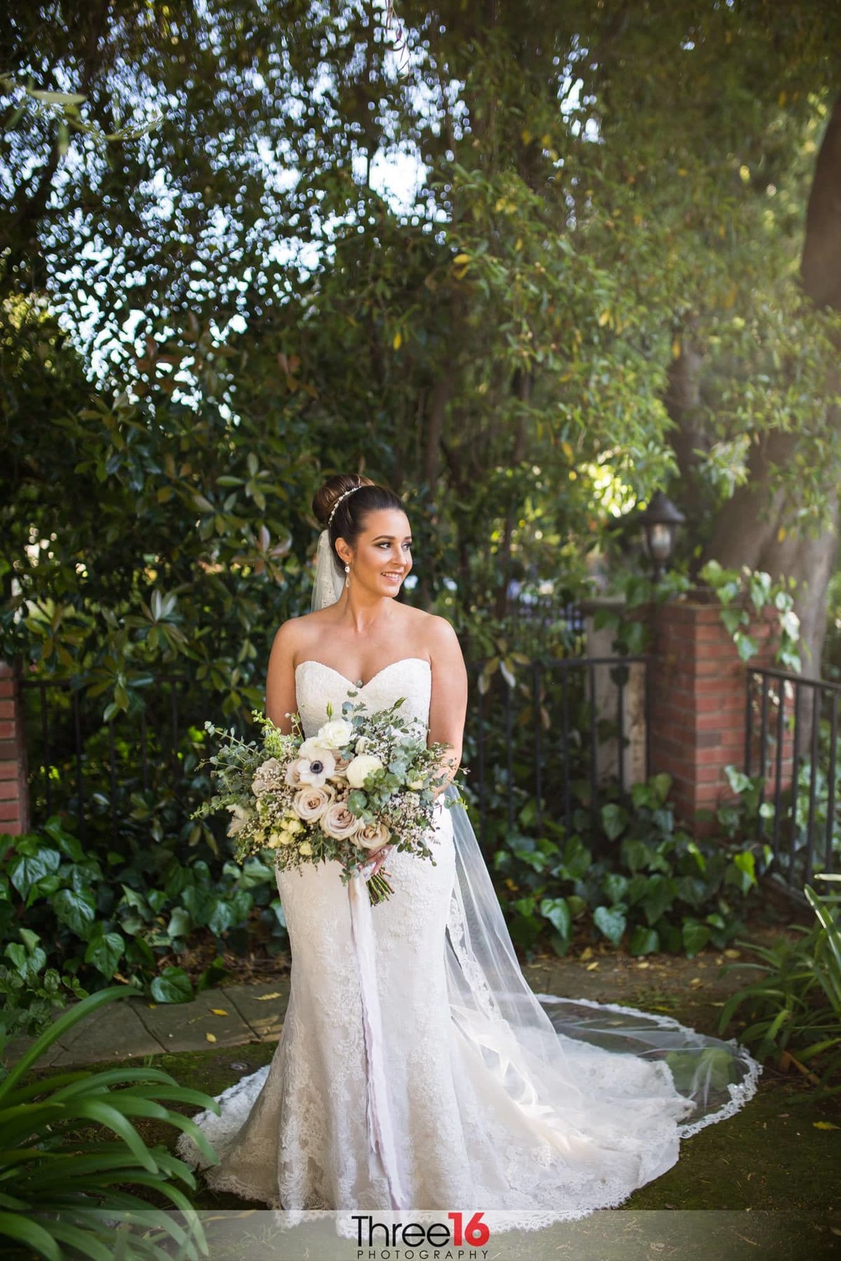 Bride poses for a photo holding her bouquet of flowers and her dress train fanned out amongst the green foliage