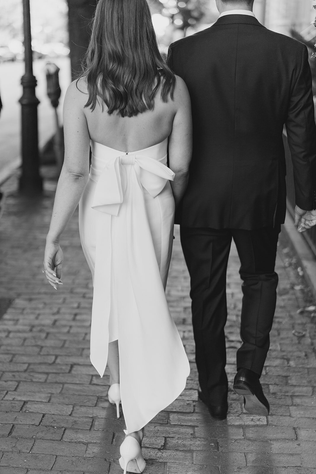Boston wedding rehearsal dinner at the Hampshire House on Boston Common. Bride and groom holding hands on the brick sidewalk to their welcome dinner. White rehearsal dinner dress with big bow on the back.