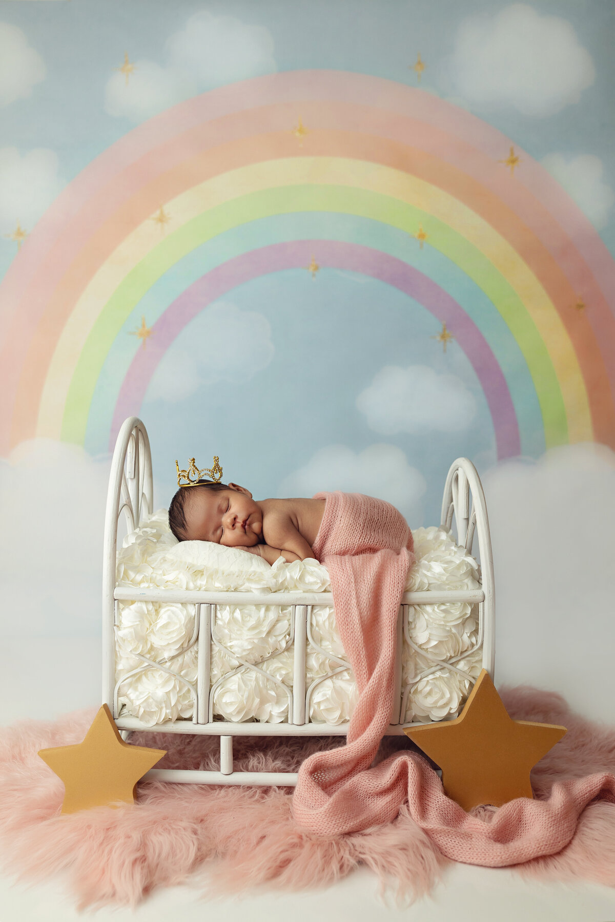 A newborn baby sleeps in a metal bed in a studio under a rainbow