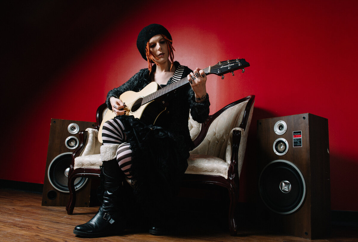 A person sitting on a couch playing an acoustic guitar with amps on either side of them.