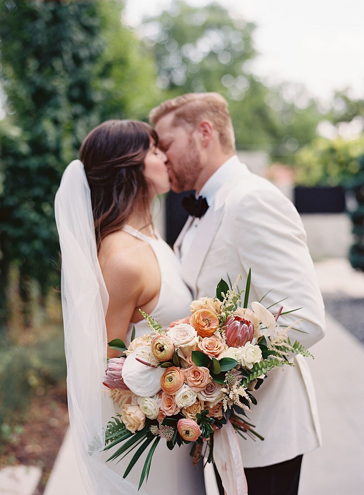 The bride, wearing a white silk halter top wedding dress and long veil kisses the groom wearing a tuxedo with a white jacket while holding her horizontal cascade bouquet of palm fronds and peach, blush, white and ivory tropical flowers.