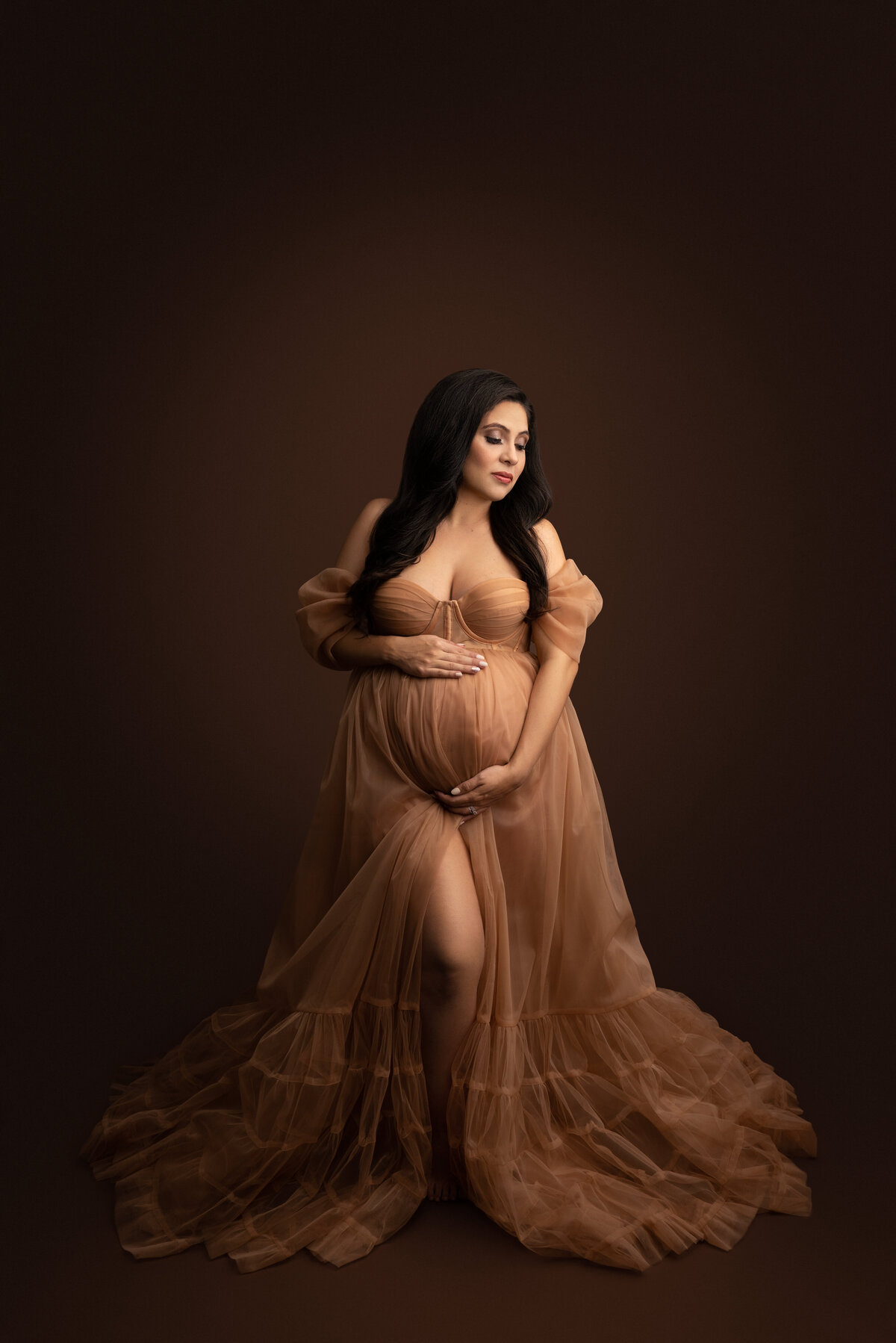 In this fine art photograph, Katie Marshall, Philadelphia's best maternity photographer, captures an expectant mom in a floor-length dark taupe organza maternity gown. The gown flows beautifully around her with a bodice resembling a corset. One hand is under her bump, the other is overtop of it. The woman is looking over her shoulder in the direction of the her elbow with her eyes closed and a peaceful expression.