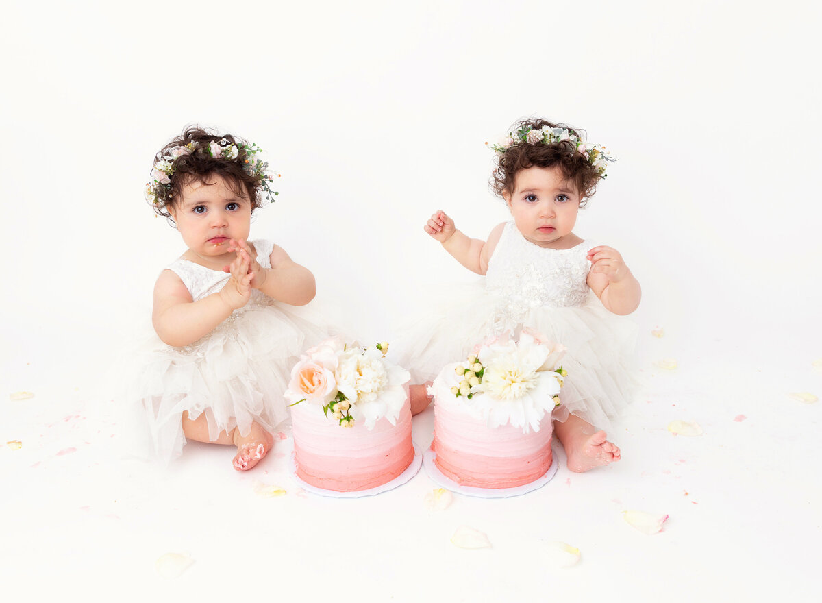 Twin baby girls in white tutus sit with pink ombre cakes between their legs.
