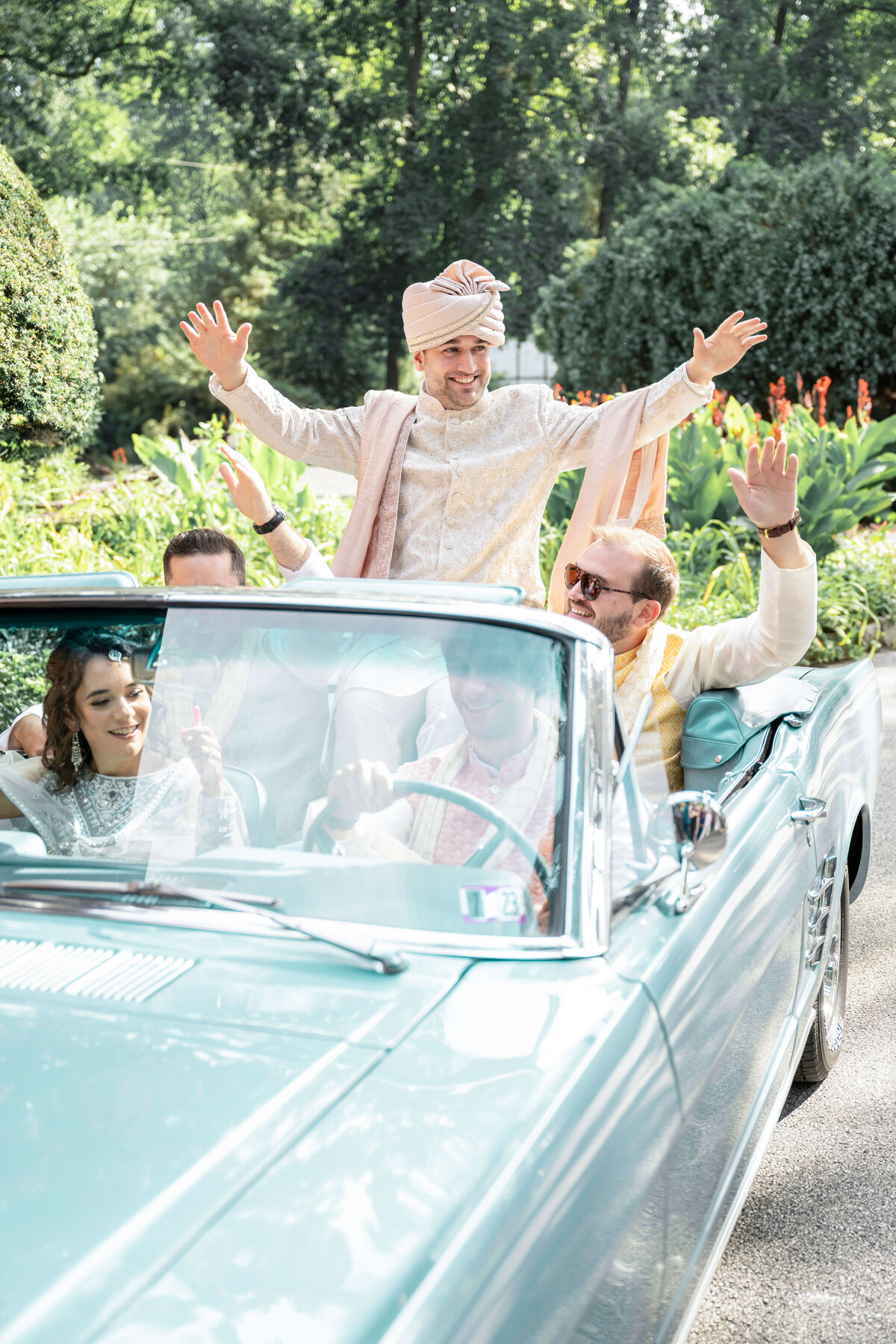 A joyful scene of people celebrating in a vintage convertible car. One man stands out joyfully with arms raised, wearing a traditional turban and outfit. Others are seated, smiling and cheering, in a lush, green garden setting.