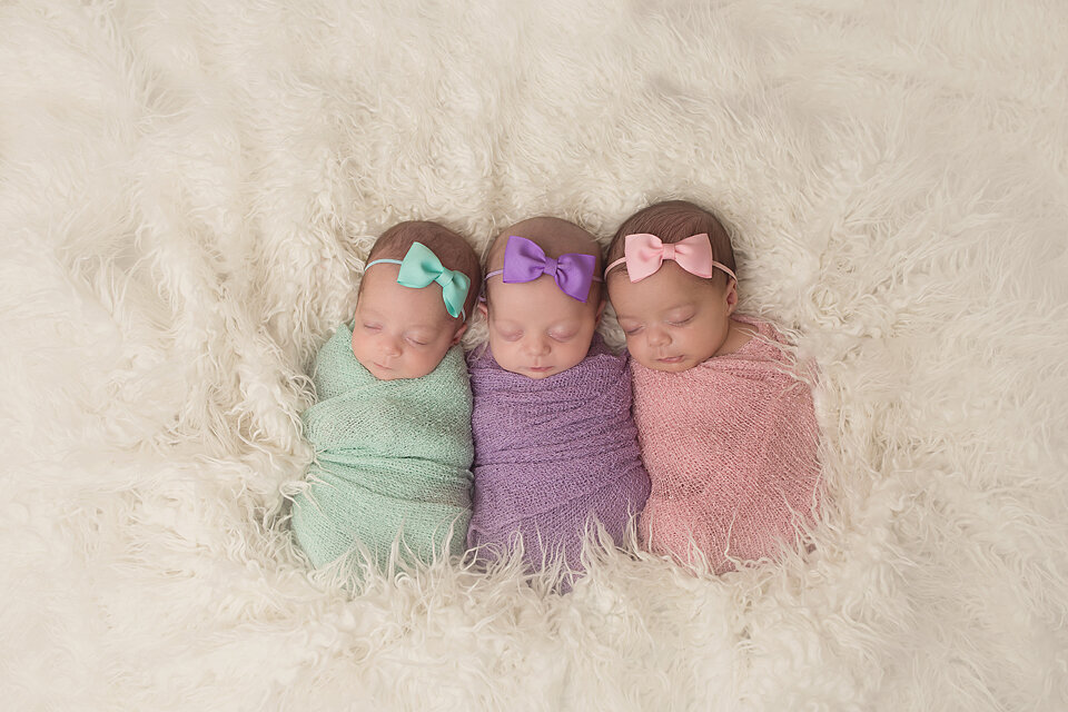 Three sleeping newborn baby girls lay in a white shag blanket in a studio in colorful swaddles and bows
