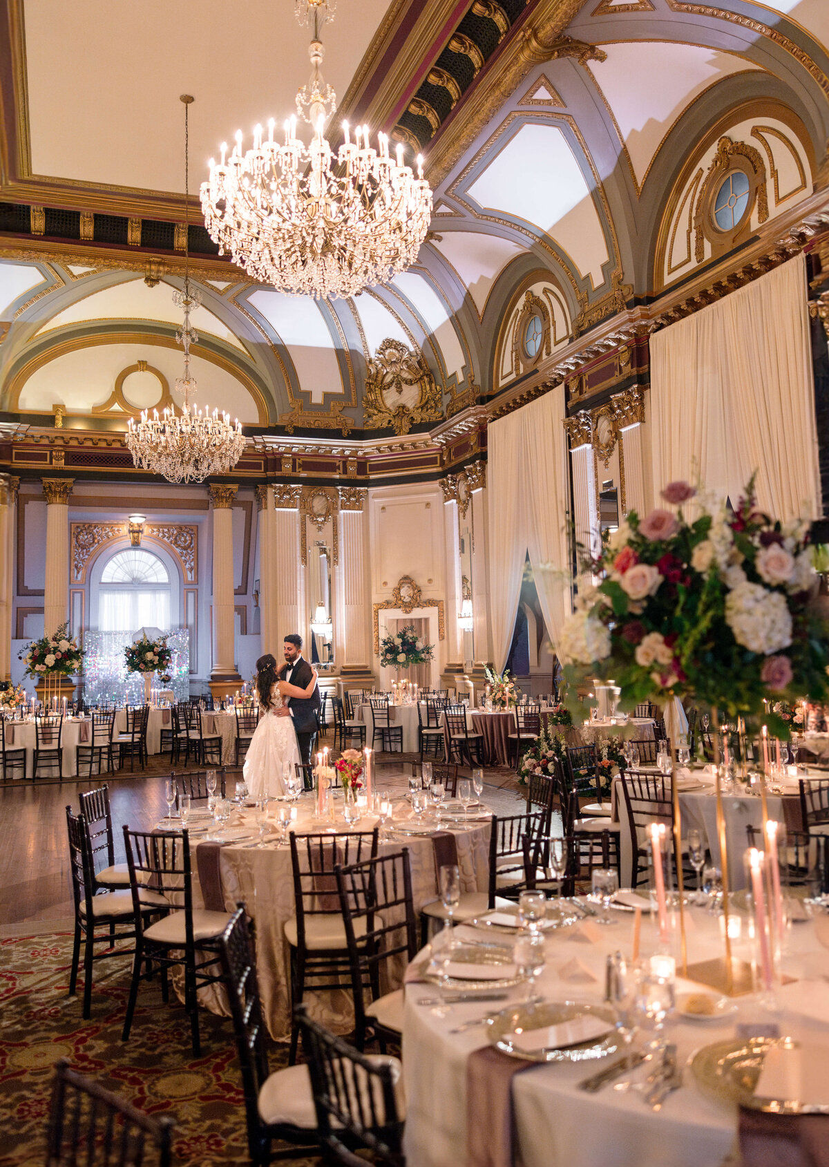 A couple dances in an elegant ballroom adorned with crystal chandeliers and ornate architectural details. Round tables are elegantly set with floral centerpieces and candles, creating a romantic and sophisticated atmosphere.