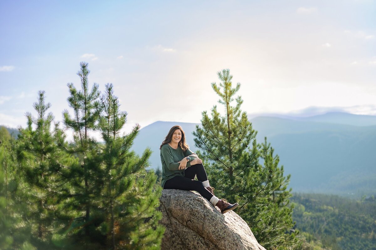 woman sitting on large rock in black leggings surrounded by pine trees and large mountains in backgroud