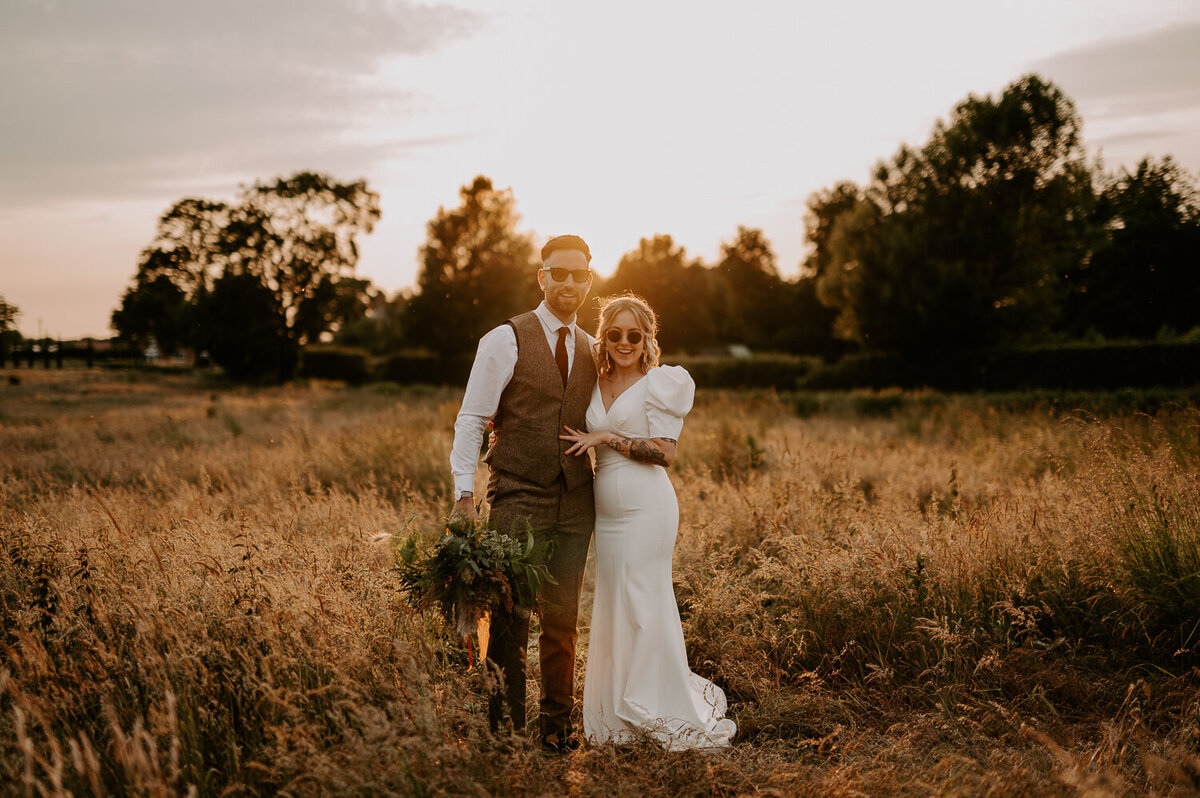 A Bride and Groom stand in the tall grass of the field outside Willow Marsh Farm with their back to an epic sunset, the bride is holding a large wild flower bouquet with pampas grass, her dress is a puffed shoulder fitted dress with a train. The Groom is wearing a brown tweed suit with burgundy tie and burgundy Dr Marten loafers.