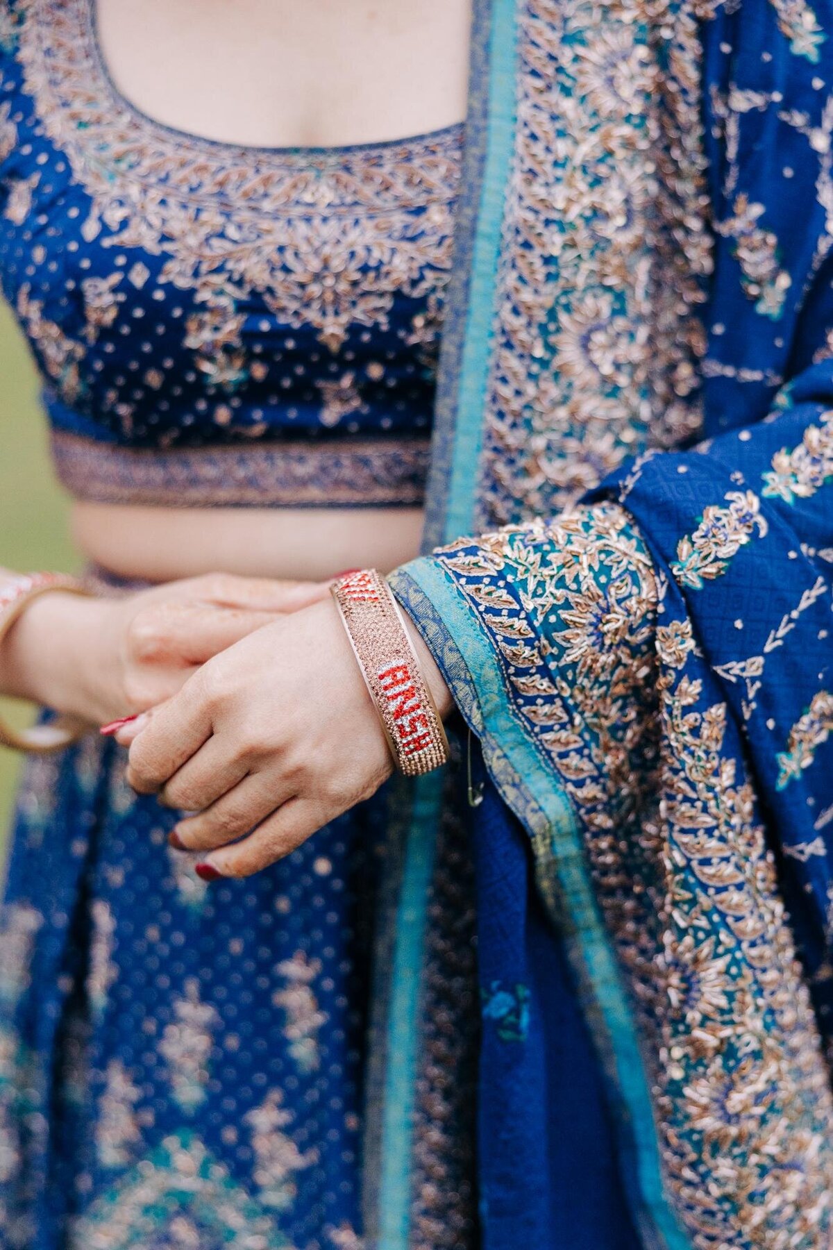 A close-up of a woman in a blue traditional embroidered dress, focusing on her hand adorned with a red and gold bracelet.