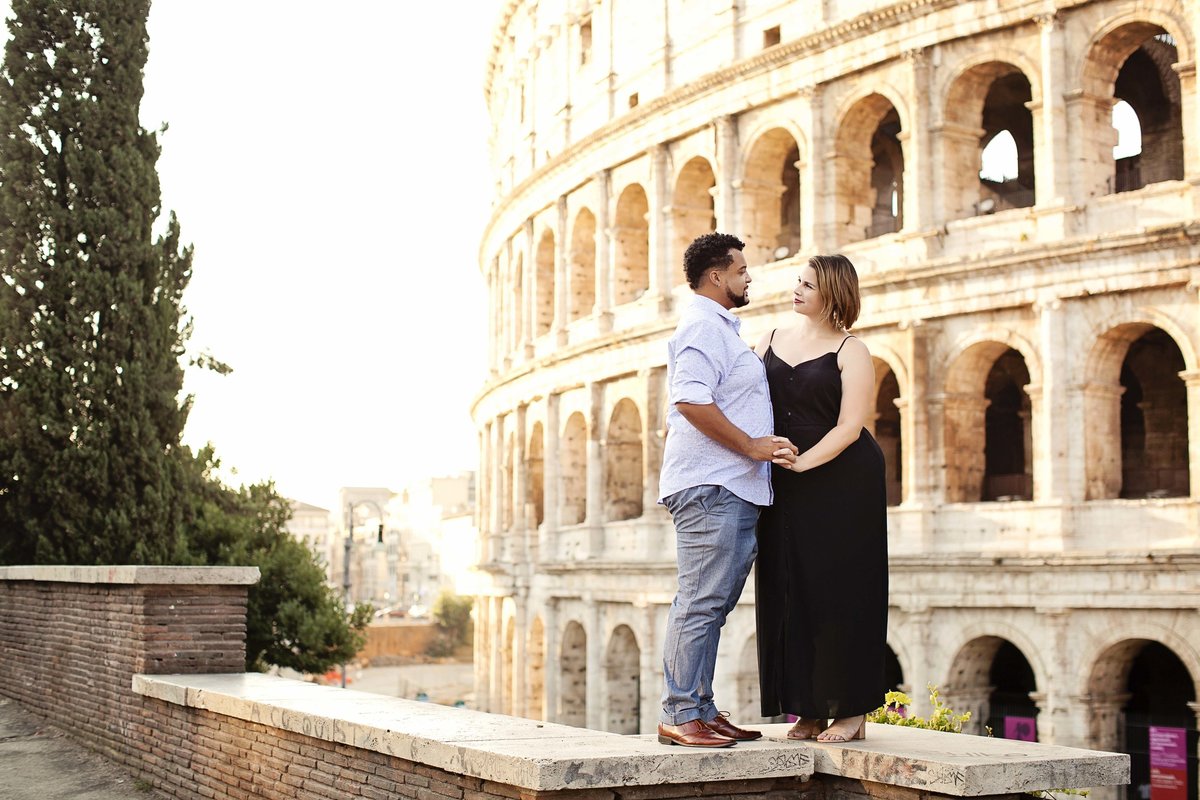 A couple on vacation in Rome standing in front of the Colosseum holding hands and looking into each others eyes. Taken by Rome Photographer, Tricia Anne Photography
