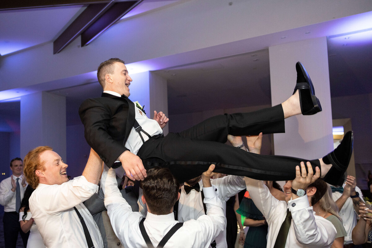 A group of people lifts a man wearing a black suit and white shirt into the air at a celebration. The setting appears to be indoors with soft purple lighting. The mood is joyful and festive.