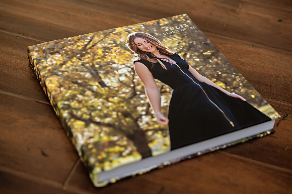 Photo Cover Session Book shown closed and laying flat on a wood backdrop, highlighting the full wraparound senior portrait cover and the smooth, durable finish of the product.