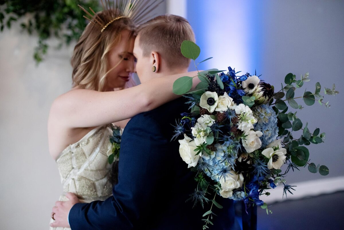 a same sex couple hugs while holding a pretty blue and white bouquet by flowers by alana