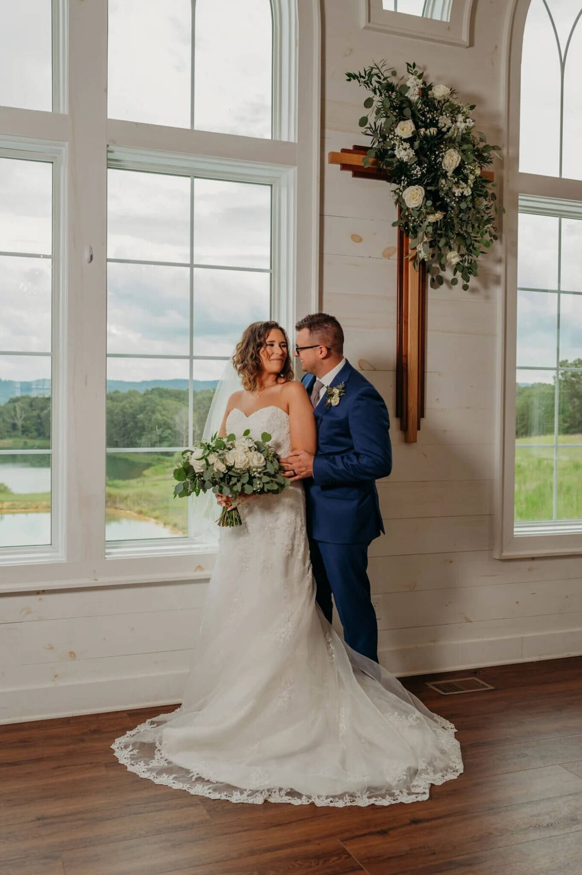 Photo of a groom hugging his bride from behind while standing beside a window with a pod and mountain showing well there is a cross hanging on the wall decorated and florals
