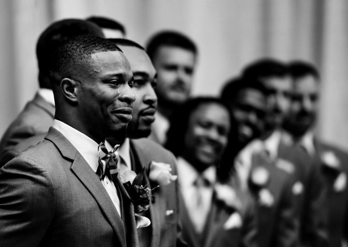 A black and white photo of a groom in a grey suit with a line of groomsmen blurred in the background.