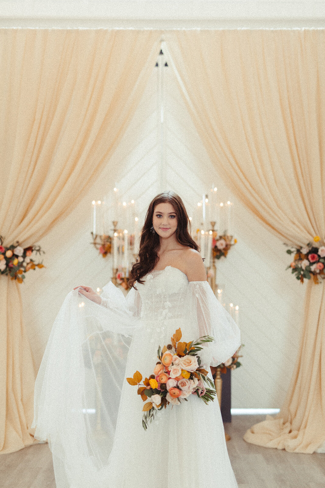 A bride wearing a white wedding gown poses with a bouquet of flowers in a bright and airy room filled with candles.