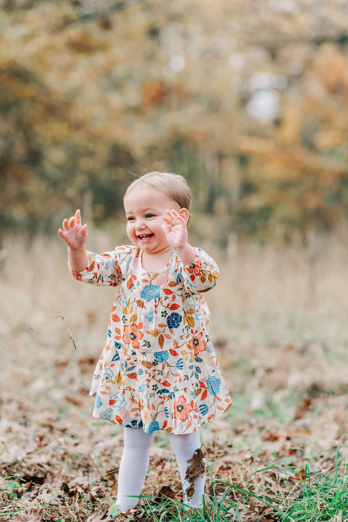 A happy, laughing  toddler wearing a floral dress holding up her hands