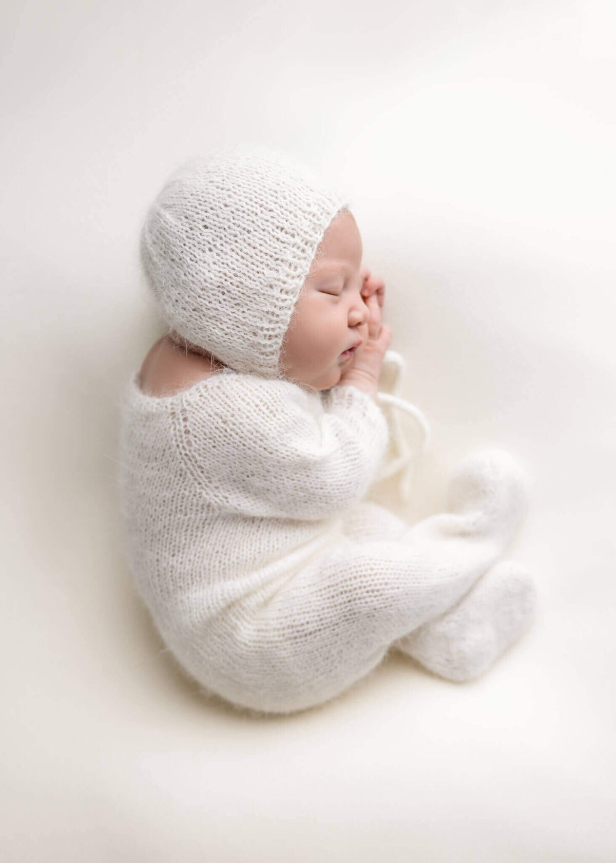 newborn baby asleep on white fabric wearing a white knitted romper and bonnet