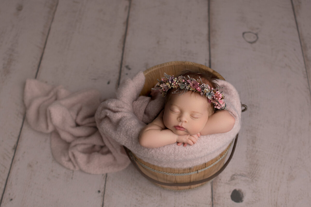 Katie Marshall, Philadelphia's top newborn photographer captures a baby girl sleeping in a bucket. Baby girls hands are folded on the bucket ledge and baby's head is resting on her forearm. Baby is wearing a large floral crown.