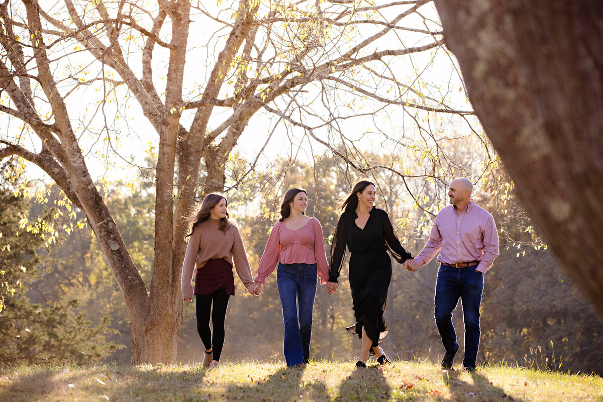 Stunning family portrait of parents and two daughters walking together on in tree‑lined field at Anne Springs Close Greenway during a sparkling November afternoon