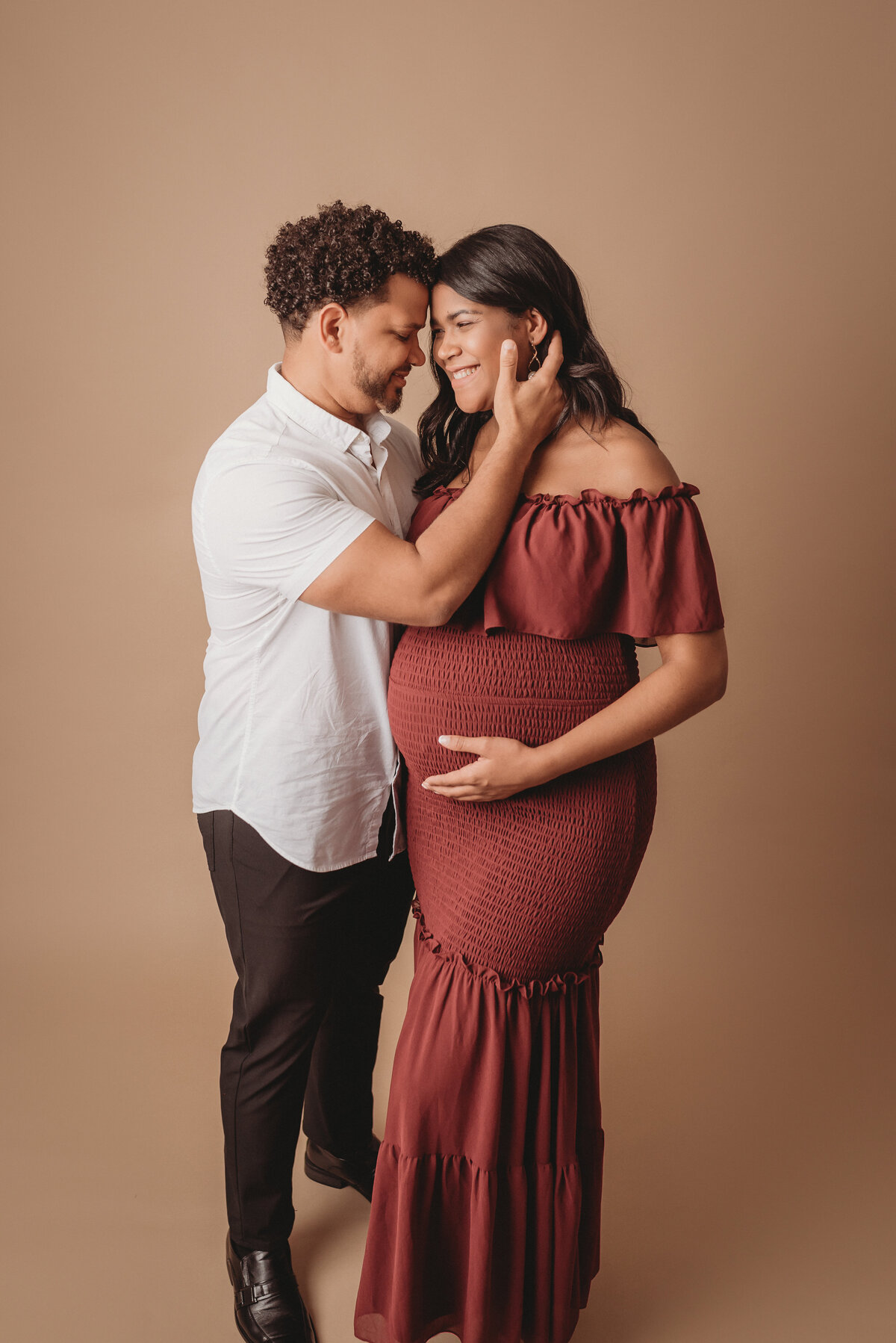 Pregnant couple standing up holding each other in maternity photo shoot wearing burgundy maternity dress with man in white button down shirt and slacks. Woman is holding baby bump.