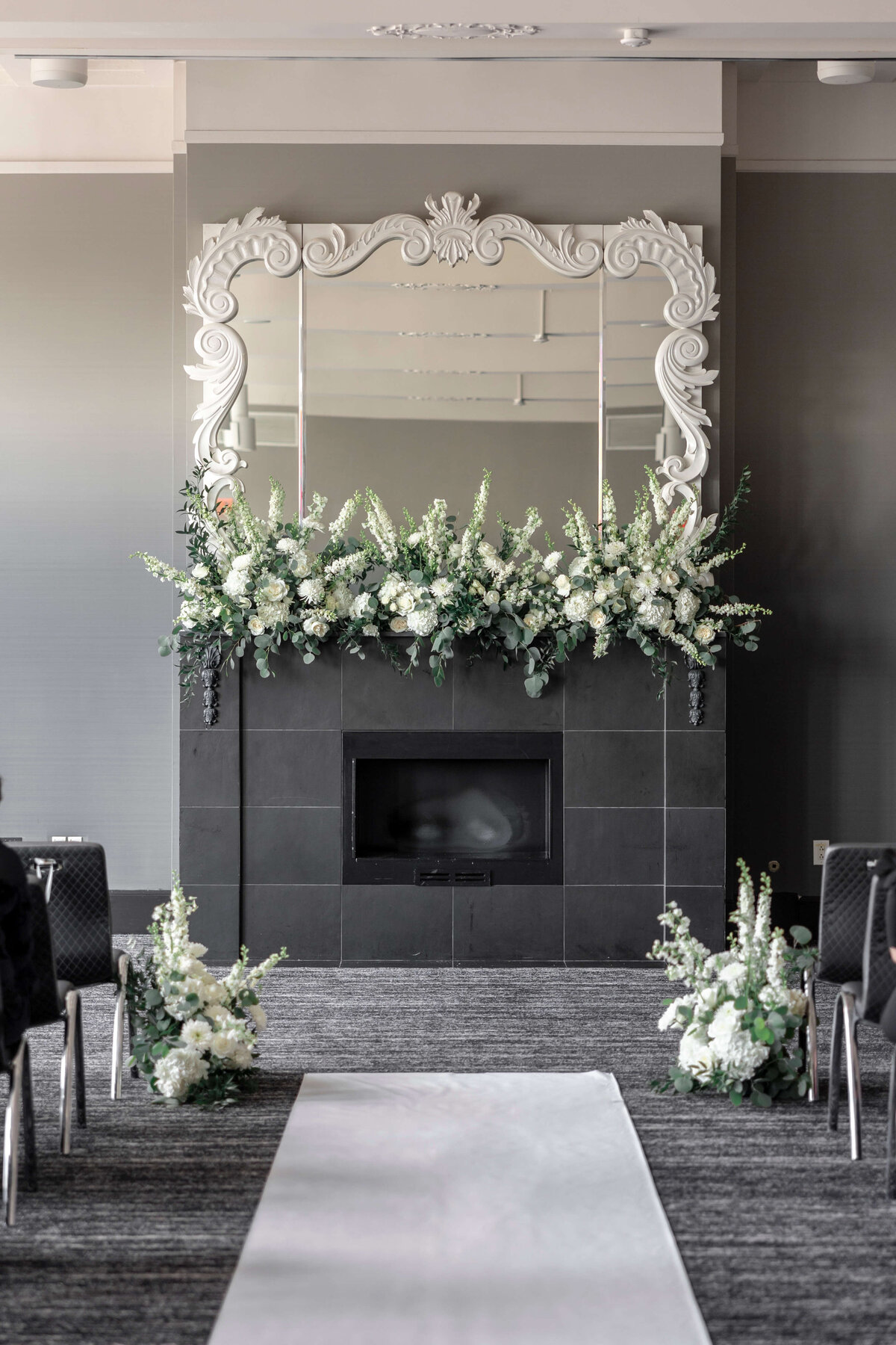 A sophisticated wedding ceremony setup features a black fireplace adorned with abundant white floral arrangements. A large ornate mirror is above the mantel, reflecting the elegant room. Rows of chairs line the aisle, which is covered with a white runner.