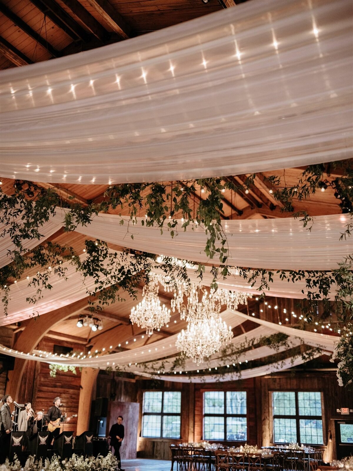 Stunning detail shot of white fabric swags hanging from ceiling alongside twinkle lights and large branches of greenery, and chandeliers at Cedar Lakes Estate wedding venue reception space.