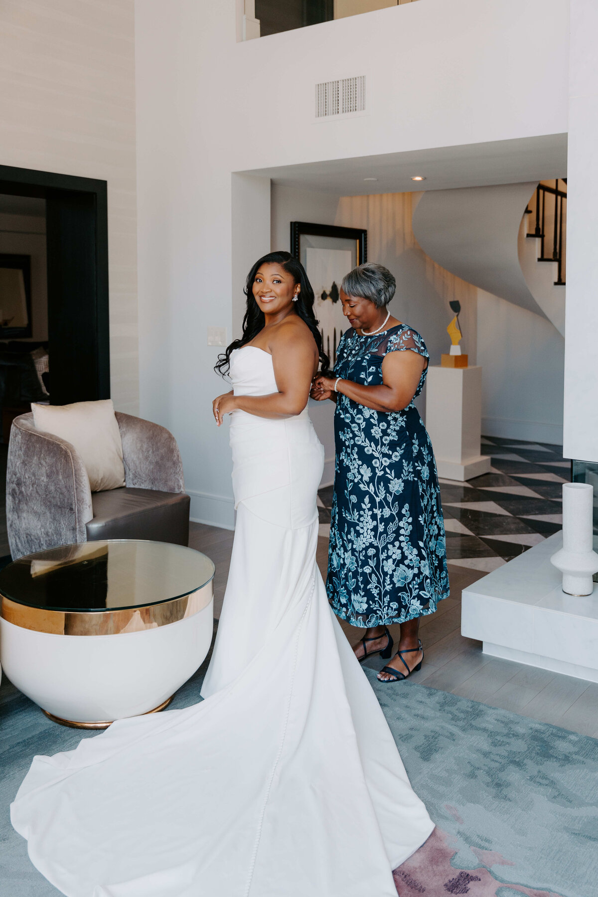 A bride in a white gown smiles warmly while an older woman in a blue floral dress fastens her dress. They are in a modern, elegant room, exuding joy and anticipation.