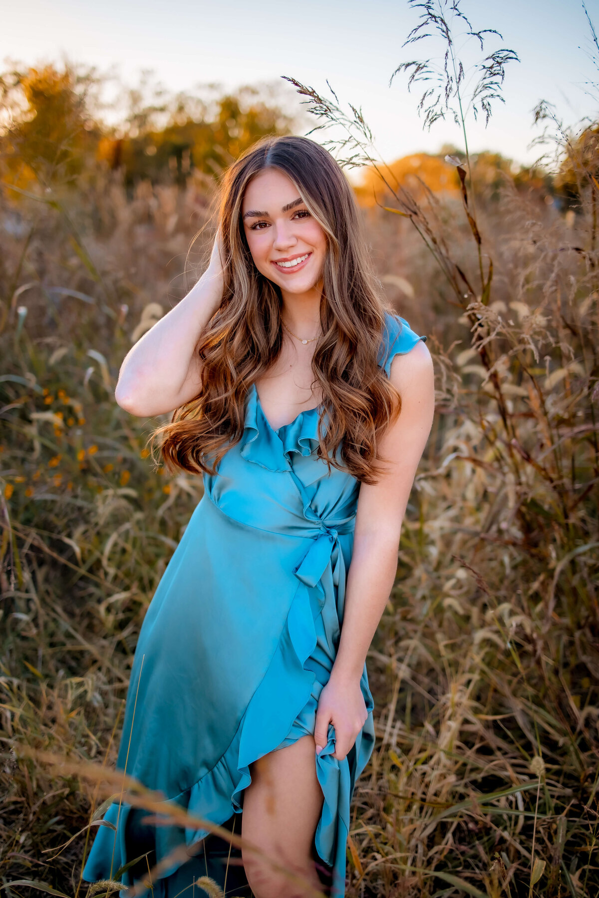 A gorgeous young lady with long brown hair is standing in a field of tall grass wearing a silky long blue dress in Clave Cave Park of St.Louis, Missouri.