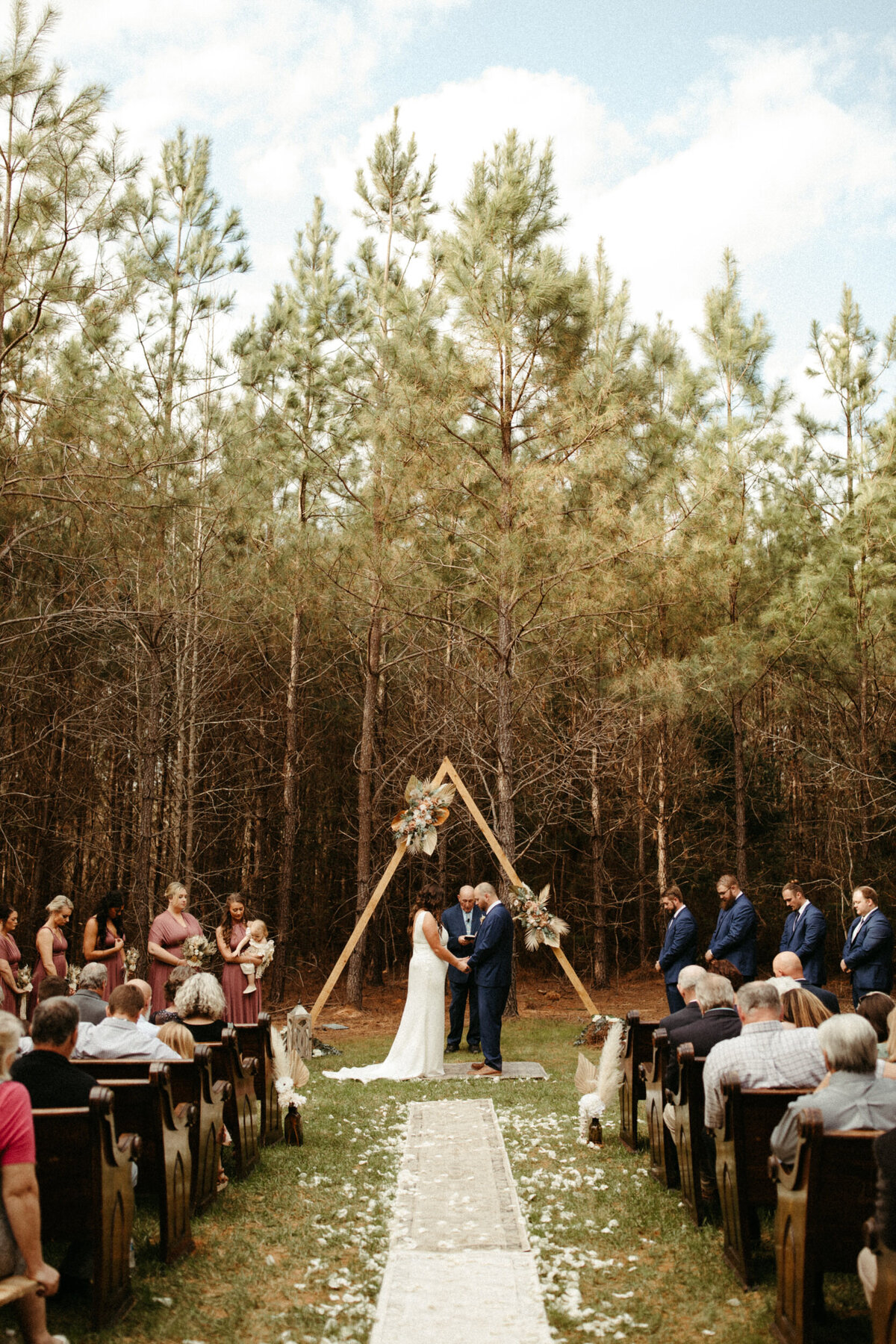 Boho wedding ceremony in the woods with vintage rugs on the aisle leading to the bride and groom standing holding hands under a triangle arch