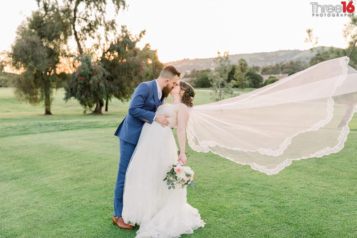 Groom dips and kisses his Bride on a golf course as her veil flies in the air