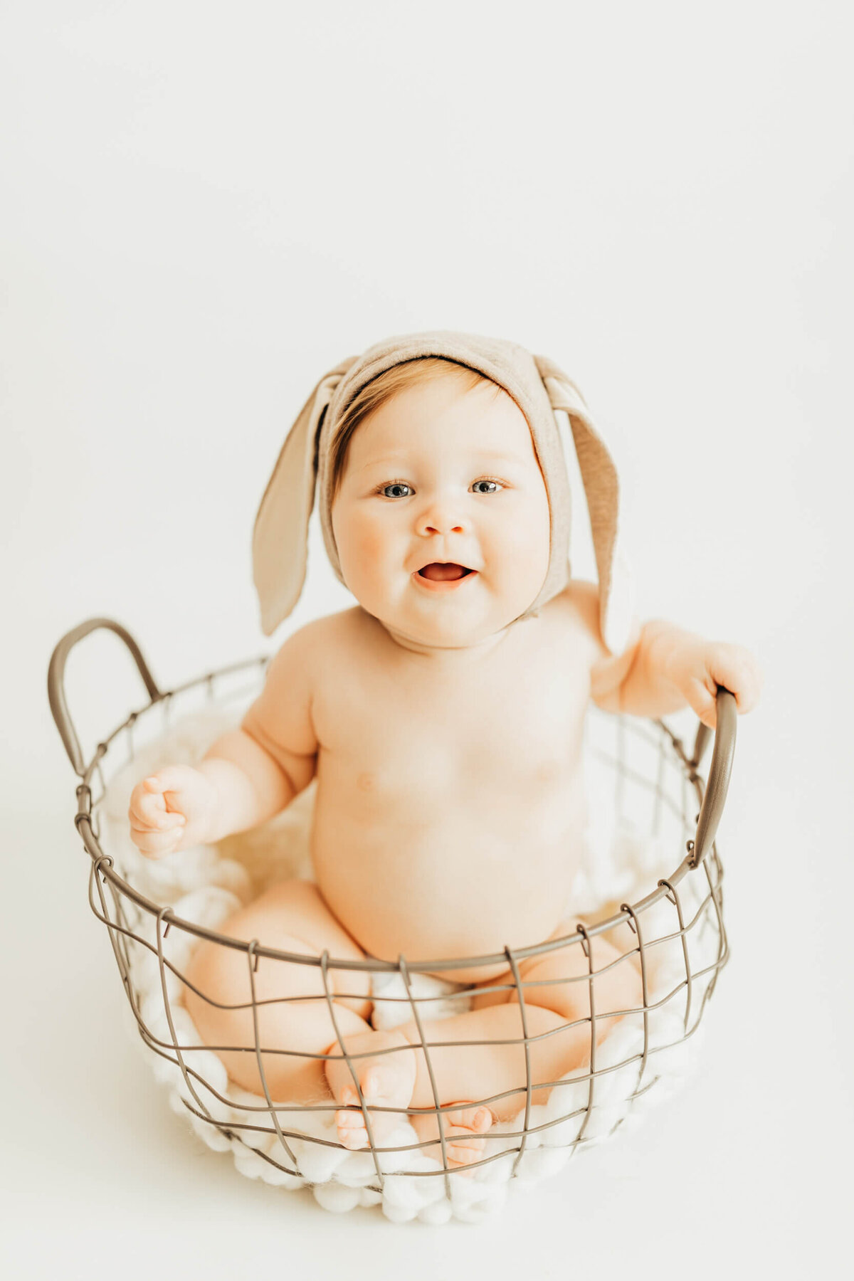 little girl wearing a bunny eared hat, sitting in wire basket on white backdrop for ally's photographyl