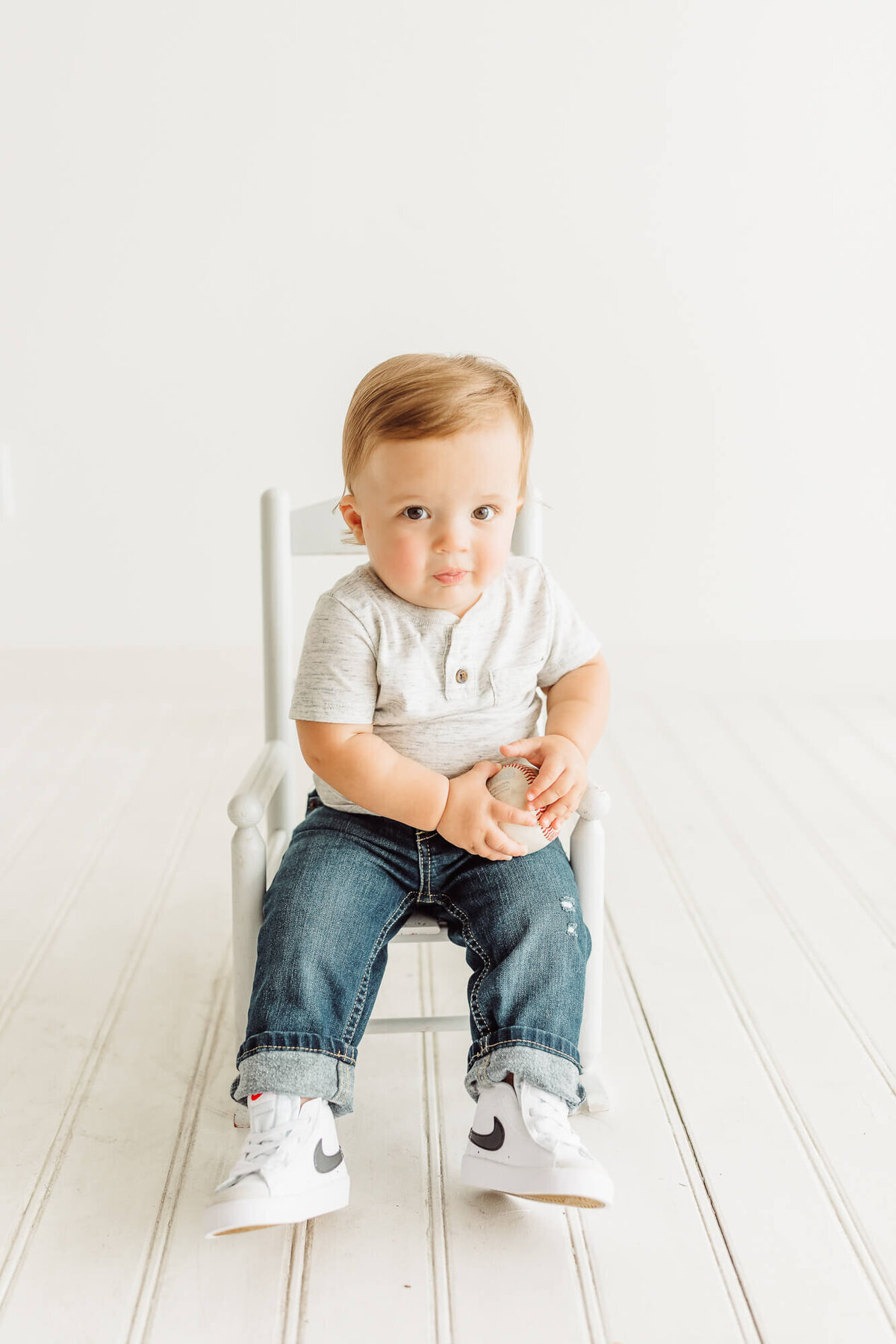 baby boy holding a baseball while sitting in white rocker in studio with Ally's Photography.