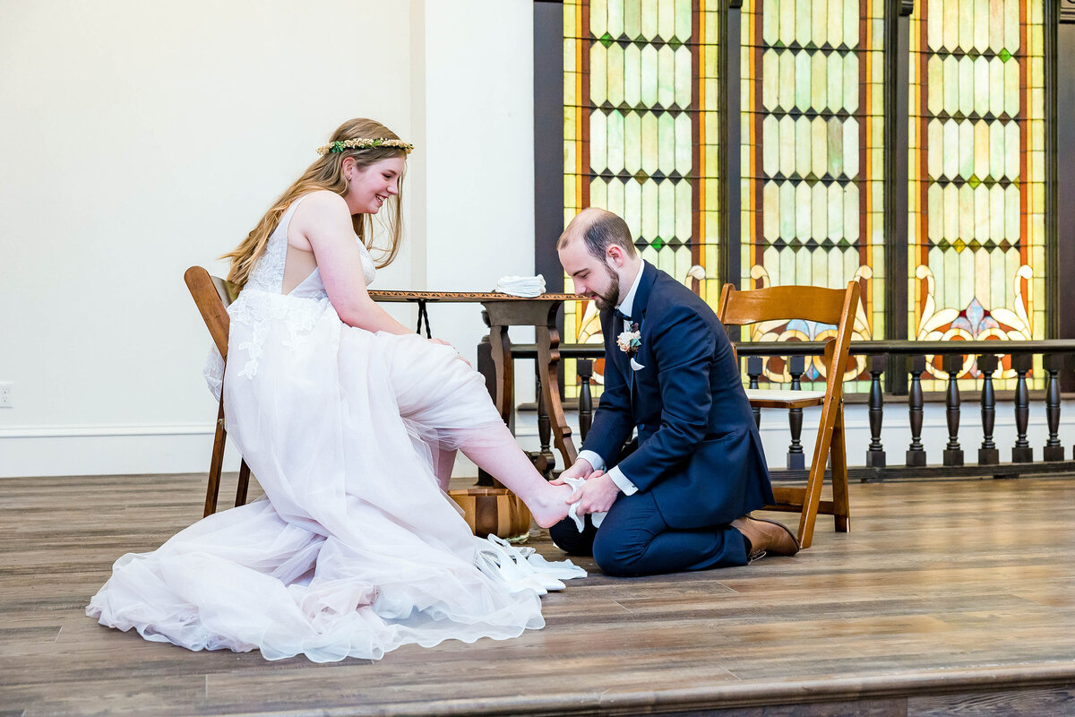 groom is wearing a blue suit and washing bride's foot during a wedding ceremony.  bride is wearing a blush lace wedding dress and watching the groom inside the chapel at Eberly Brooks Event.