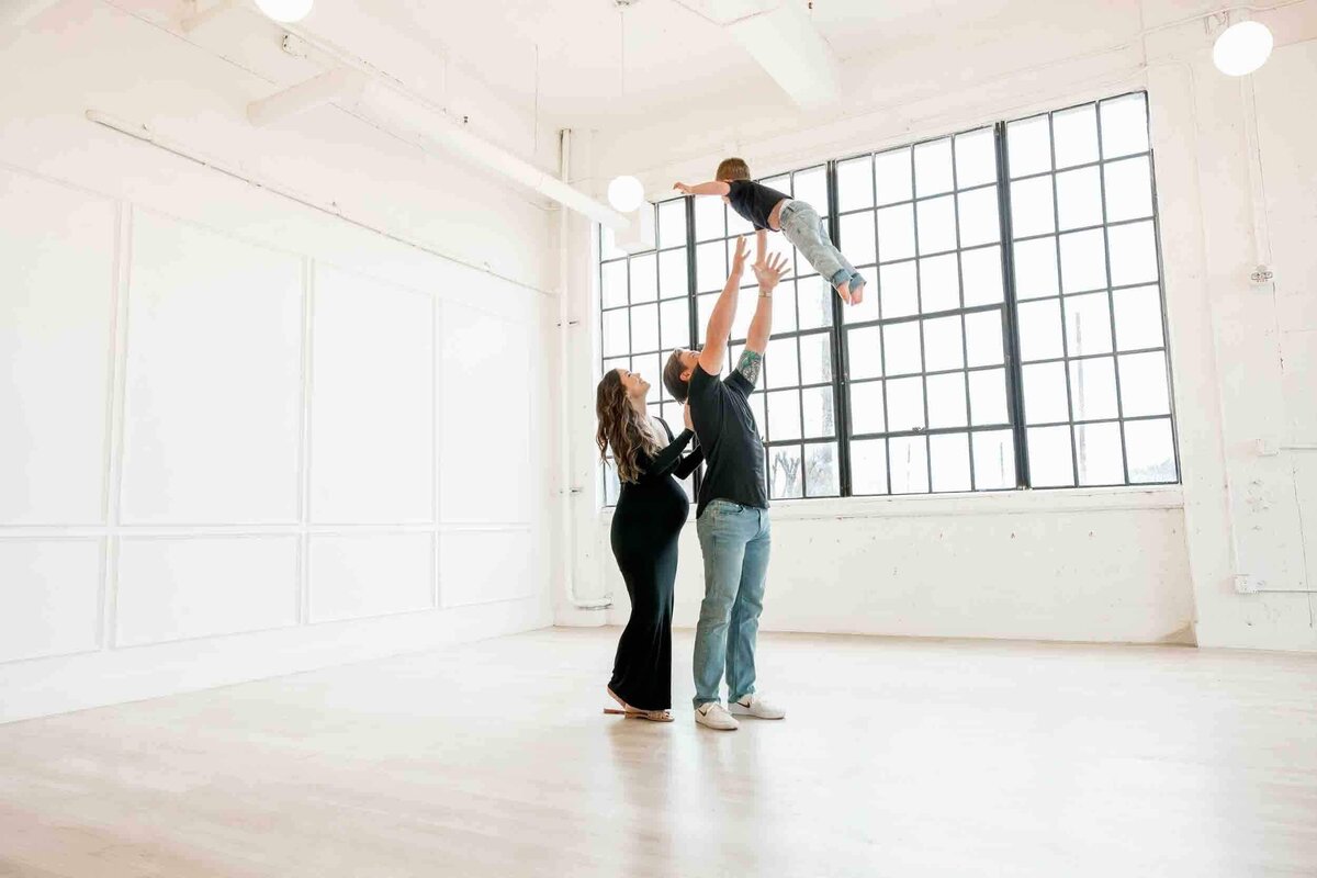 father tosses son into air in studio session while mom watches over dads shoulder, wearing black maternity dress.