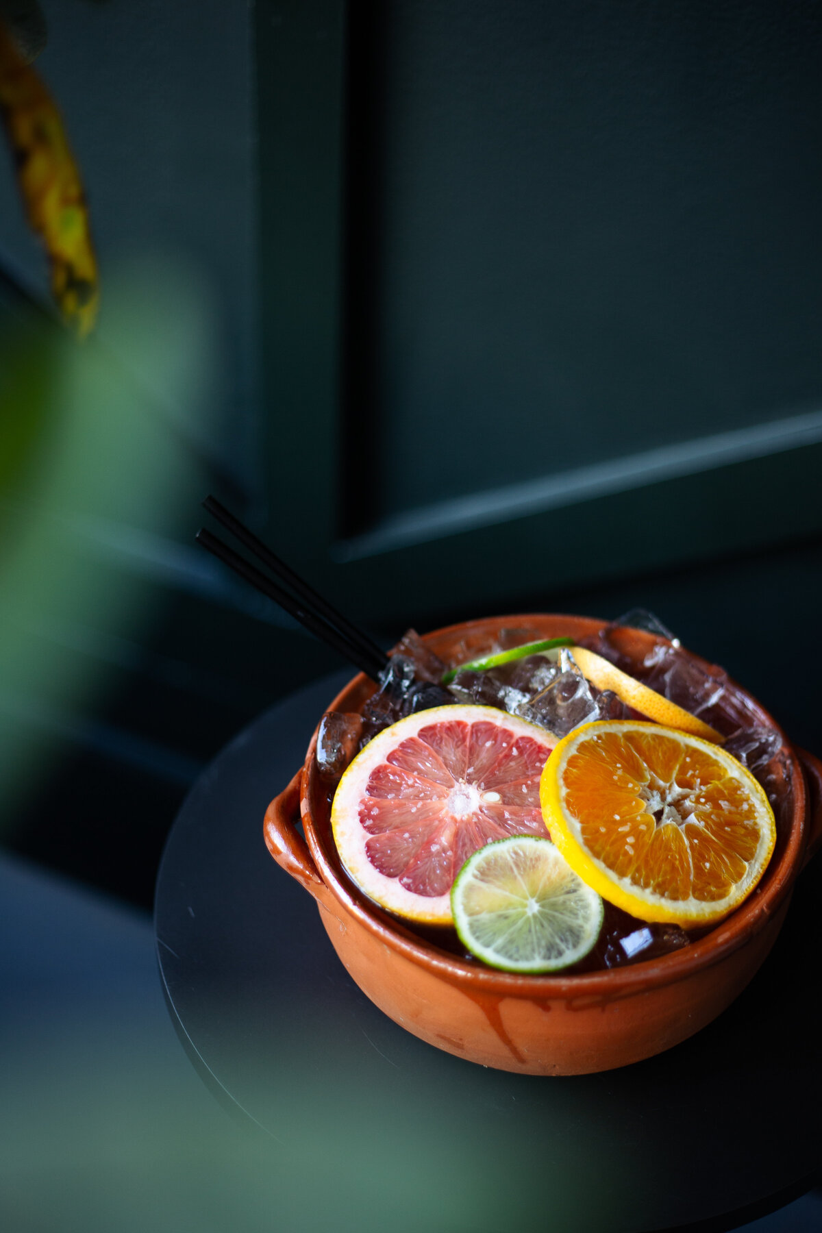 A cocktail containing fresh fruit slices photographed through leaves sits on a table in front of a dark green wall.
