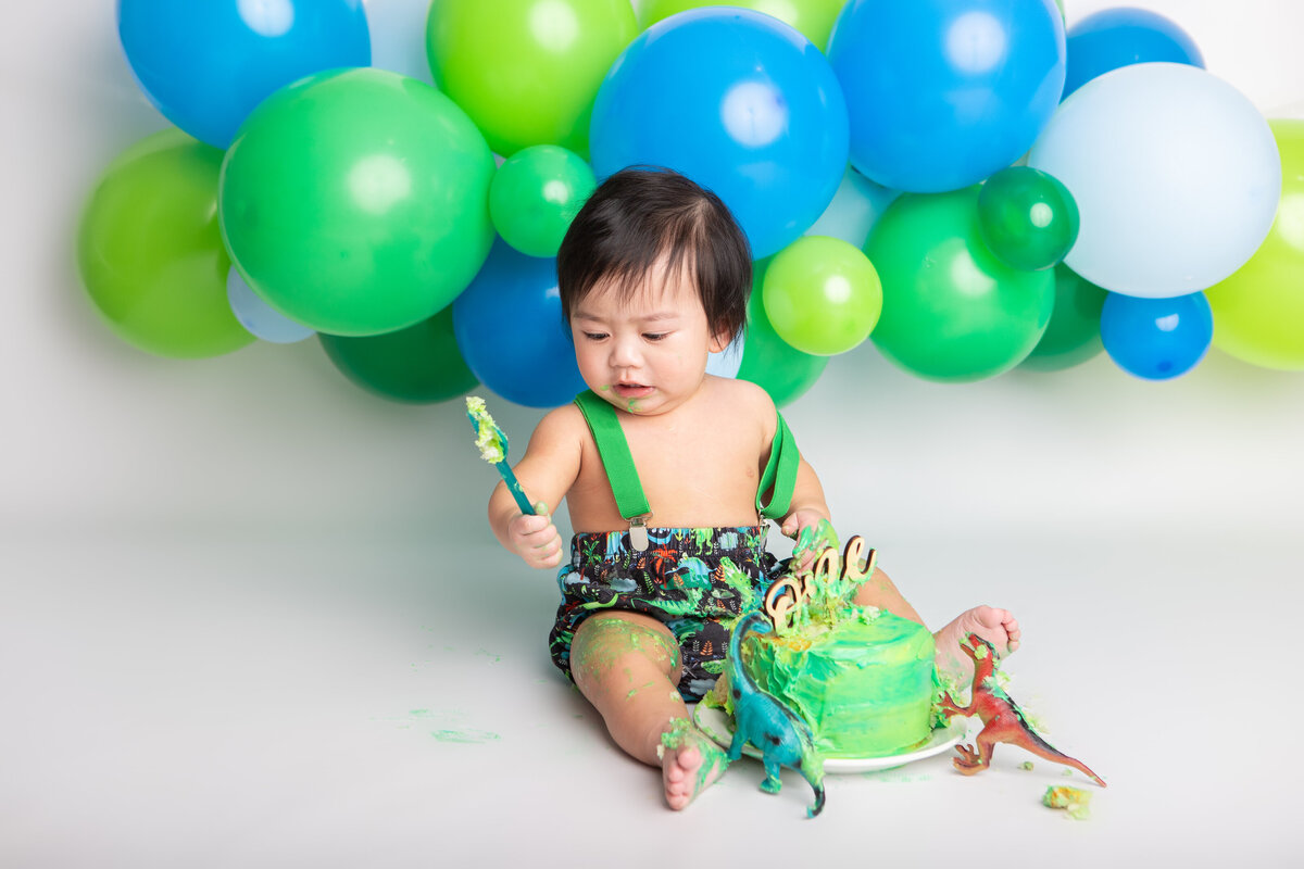 First birthday boy eating a green cake with dinosaurs and a green blue and white balloon garland
