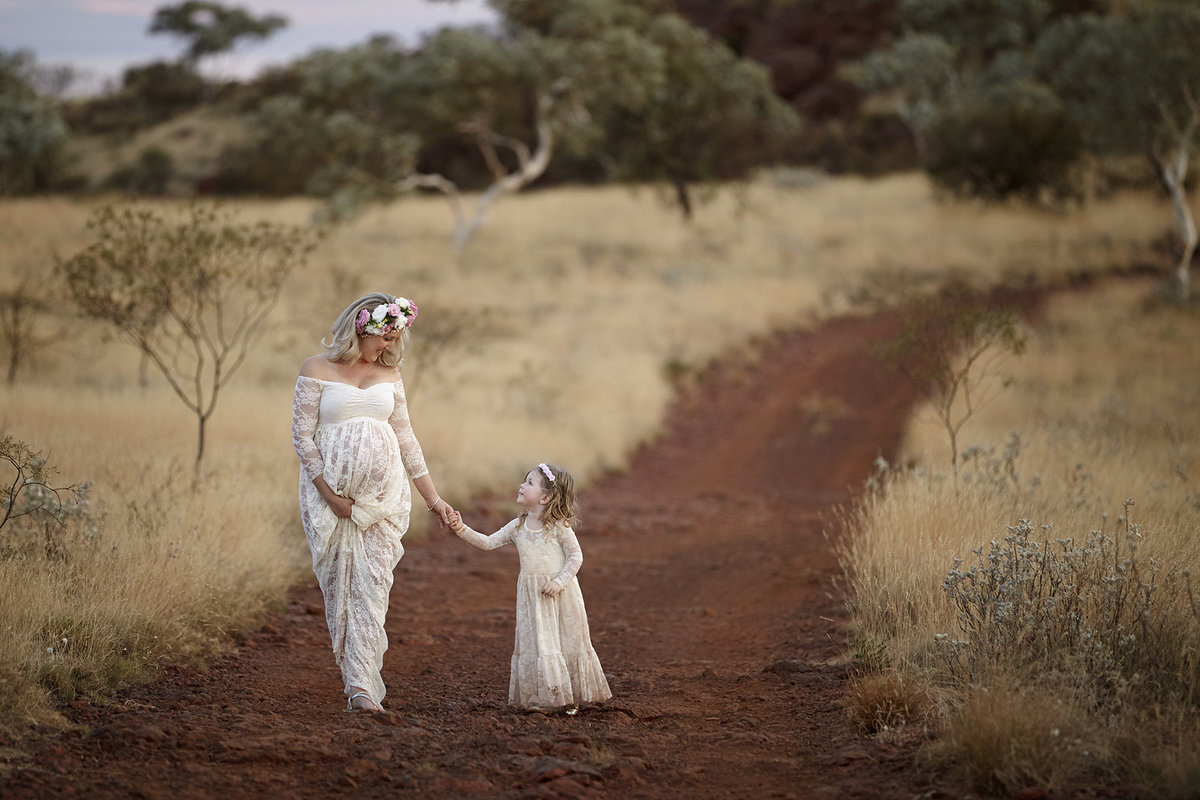 Pregnant mum holding daughters hand walking down a red dirt path looking at each other in matching white outfits