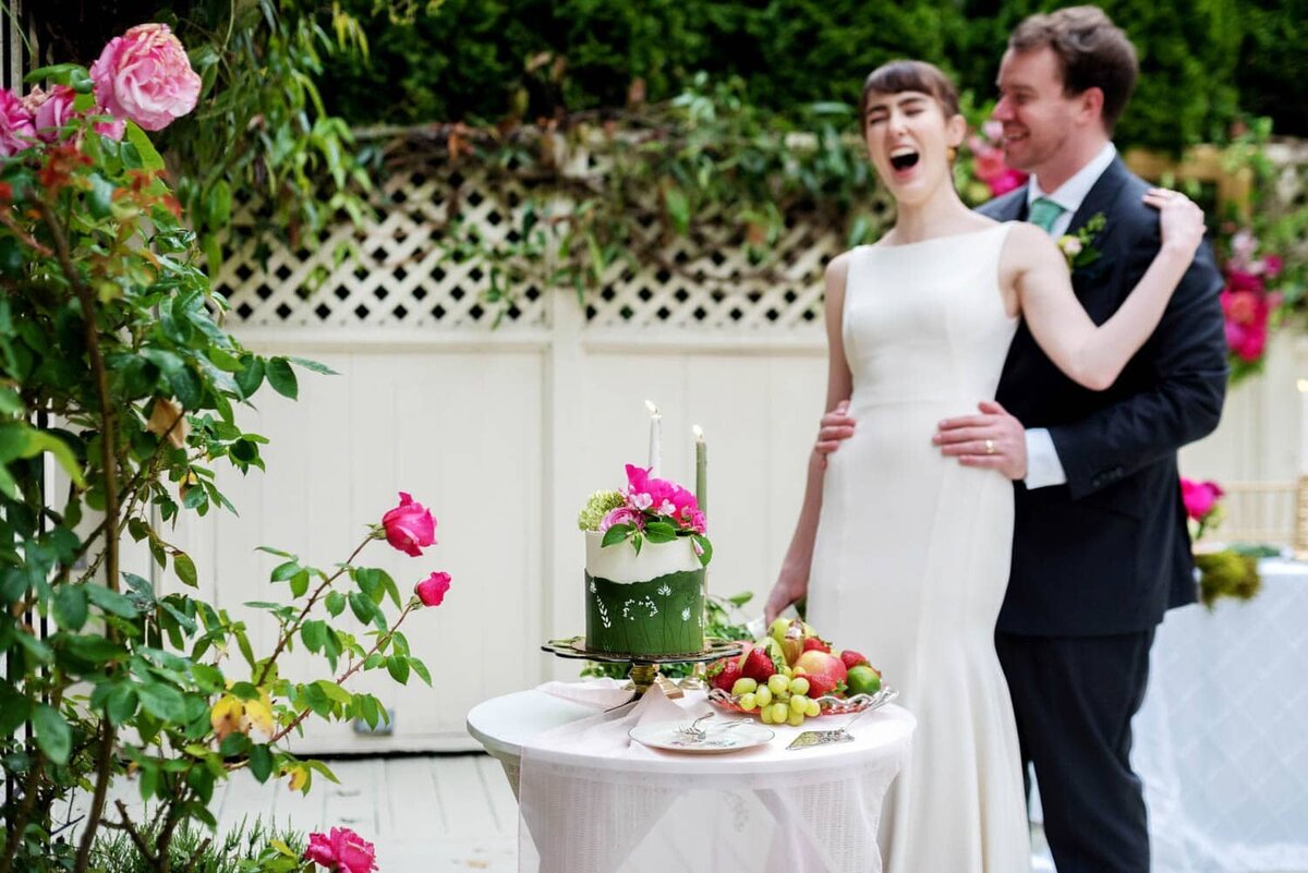a bride laughs hard and leans on the groom in front of a pretty green and white wedding cake and table of fruit