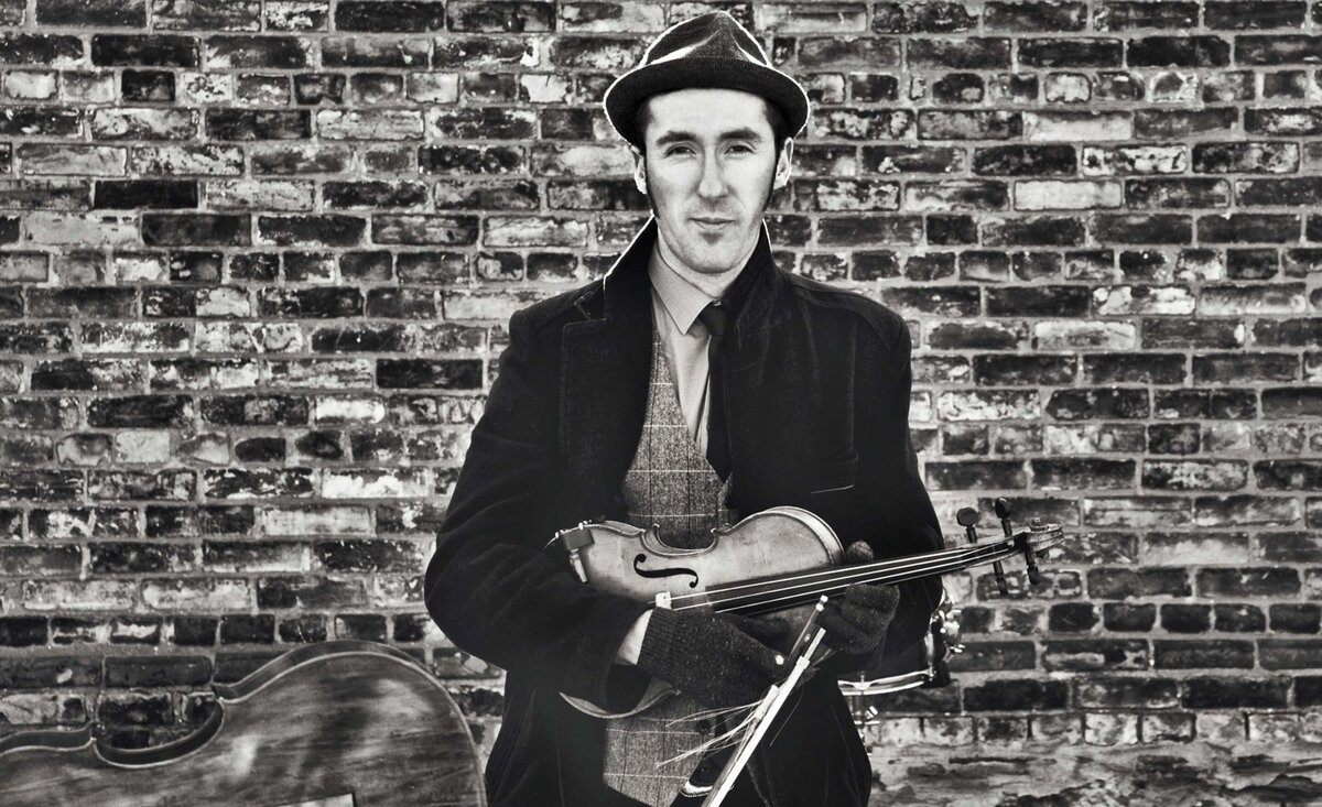 Classical music photo black and white Gordie MacKeeman wearing black coat black fedora holding fiddle against brick wall background