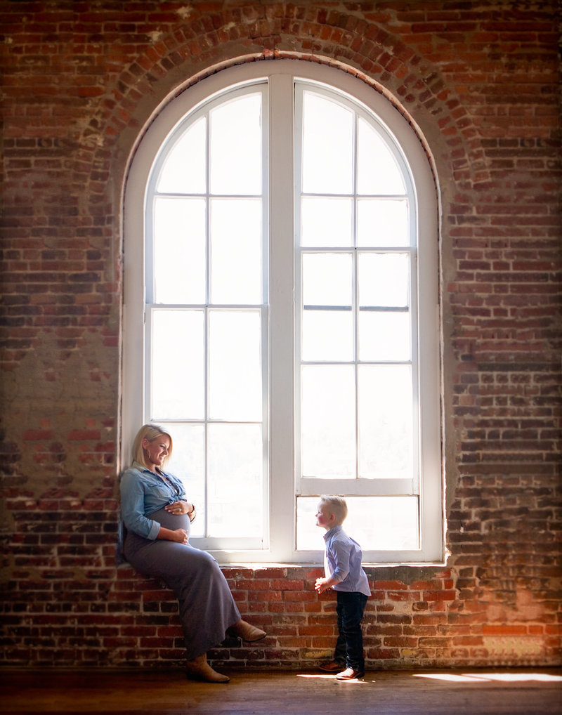 Sky 9 Studio | Professional family maternity photo of mother and son in front of large round top window against a brick wall.  Mother is pregnant and her son is lovingly looking up at her