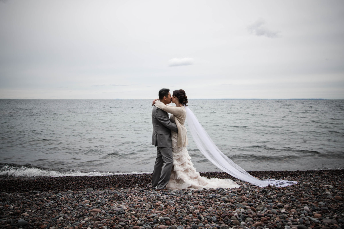 Bride and groom kiss in front of Lake Superior on cloudy day.