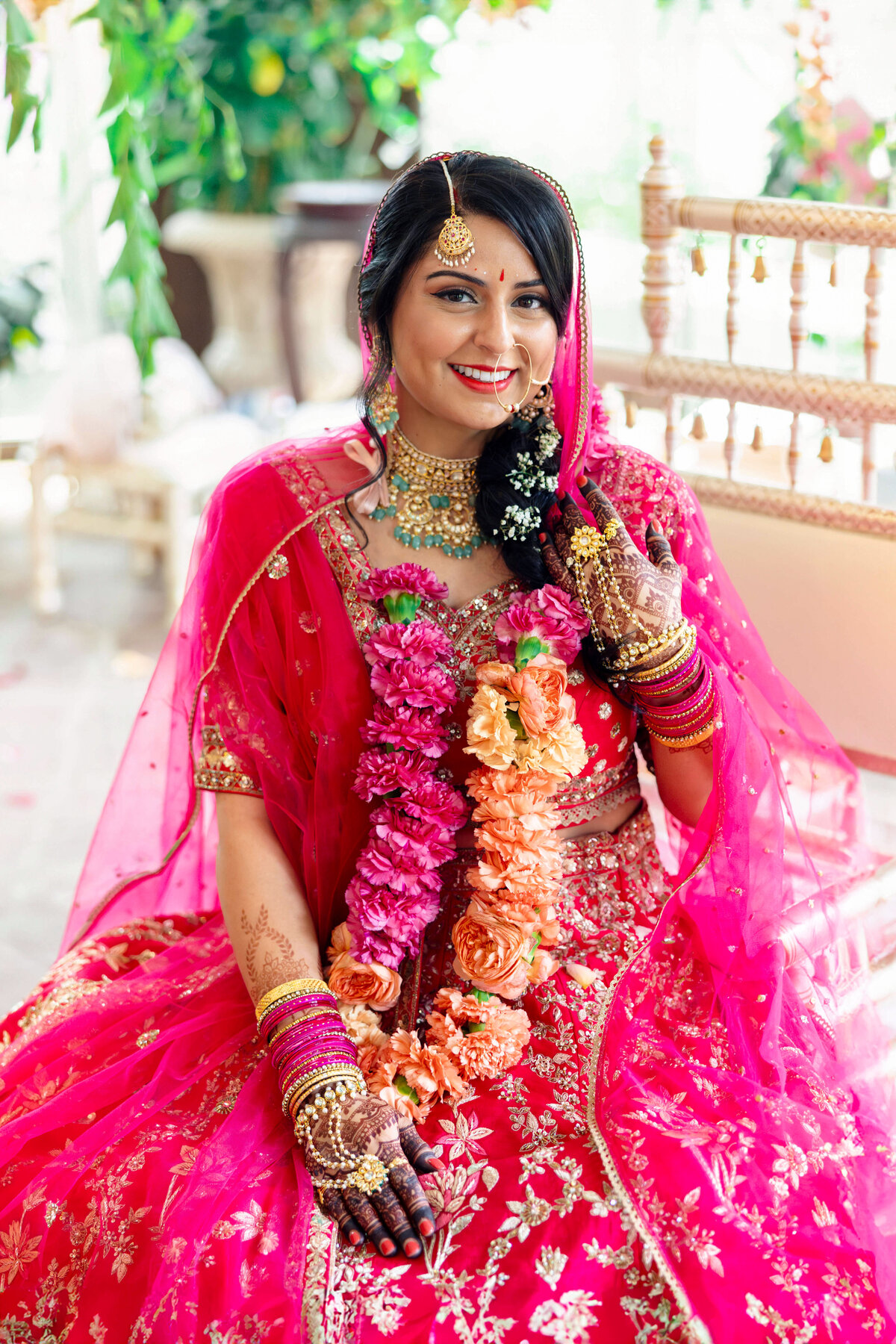 A woman in a vibrant pink and gold traditional outfit sits smiling. She wears floral garlands, intricate jewelry, and henna on her hands. The setting is adorned with greenery and elegant furniture.