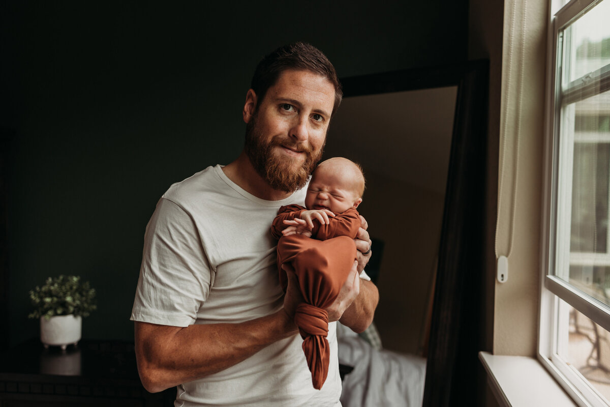 dad holds new baby boy near window wearing a rust colored sleeper sack